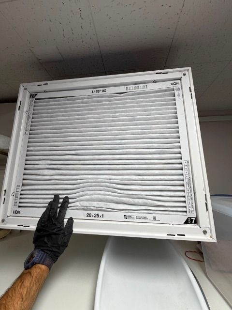 Gloved hand holding a pleated air filter beneath a ceiling return grille